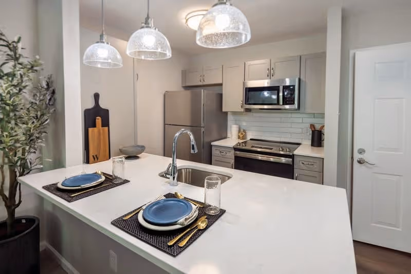 Modern kitchen with a white countertop island featuring two place settings with blue plates, gold utensils, and clear glasses. The kitchen includes stainless steel appliances such as a refrigerator, microwave, and oven. There are three pendant lights hanging above the island, a potted plant to the left, and a white door on the right side.