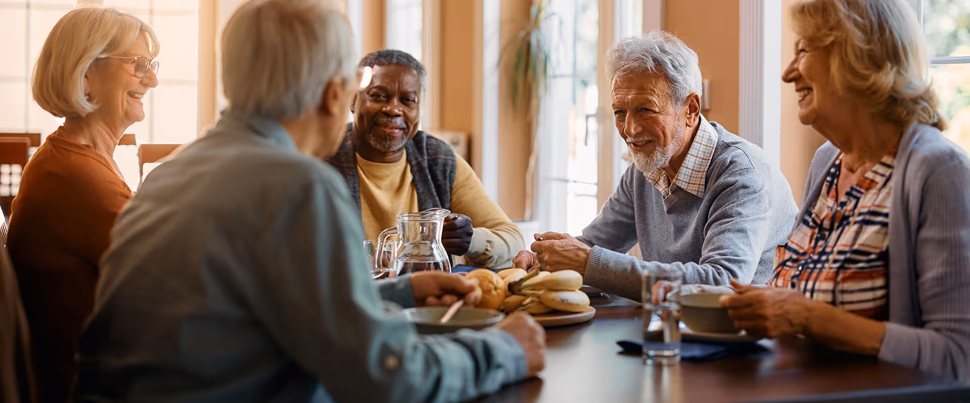 A group of five elderly people sitting around a dining table, smiling and engaging in conversation. The table has a plate of bagels, a glass pitcher of water, and bowls in front of some individuals. The setting is bright with natural light coming through windows in the background.