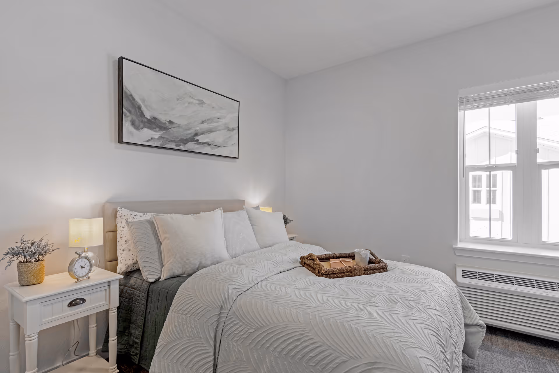 A neatly made bed with white and light gray bedding in a bright bedroom. There is a white nightstand with a small lamp, a clock, and a potted plant on the left side of the bed. Above the bed hangs a rectangular abstract painting in shades of gray. A window with blinds is on the right side, allowing natural light into the room.
