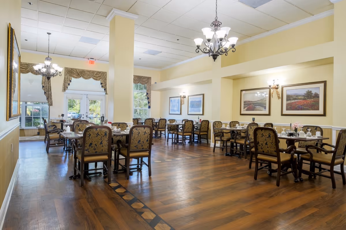 Spacious, well-lit dining room with multiple tables and upholstered chairs, chandeliers, and framed artwork on the walls.