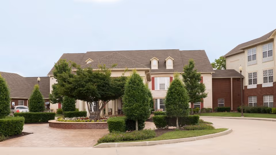 Front exterior of a multi-story assisted living building with a circular driveway, landscaped island, and trimmed trees.
