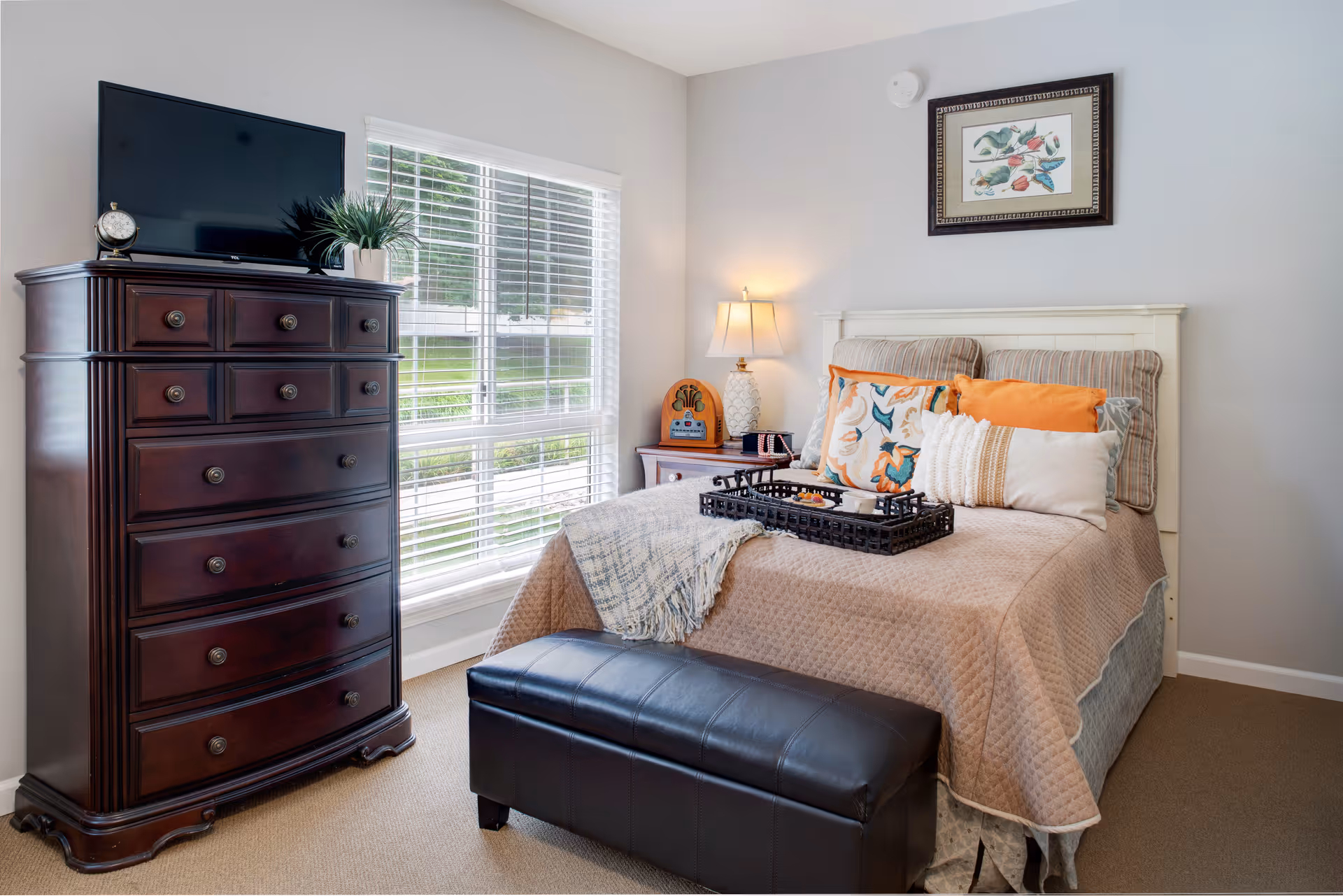 A neatly furnished bedroom with a bed topped with decorative pillows, a dark wood dresser with a TV, and a window with blinds.