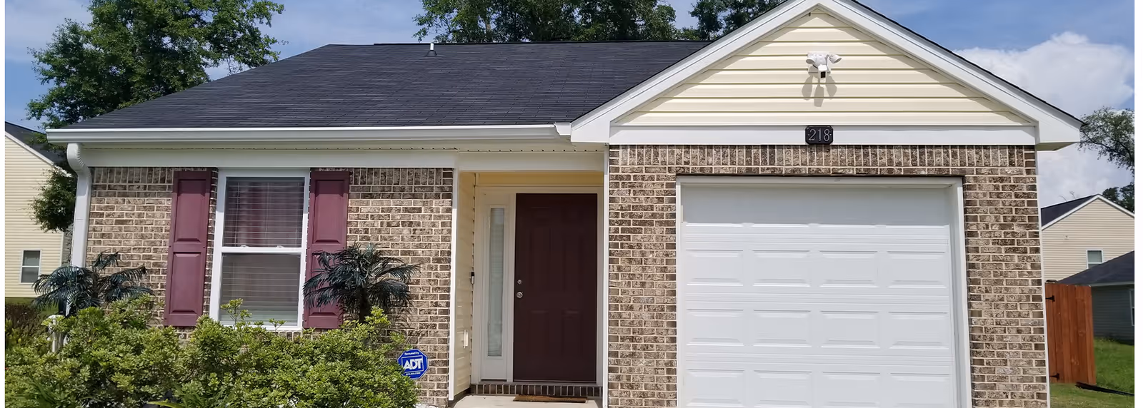 Front exterior of a single-story brick house with a white garage door, maroon front door and shutters, and shrubs in front.