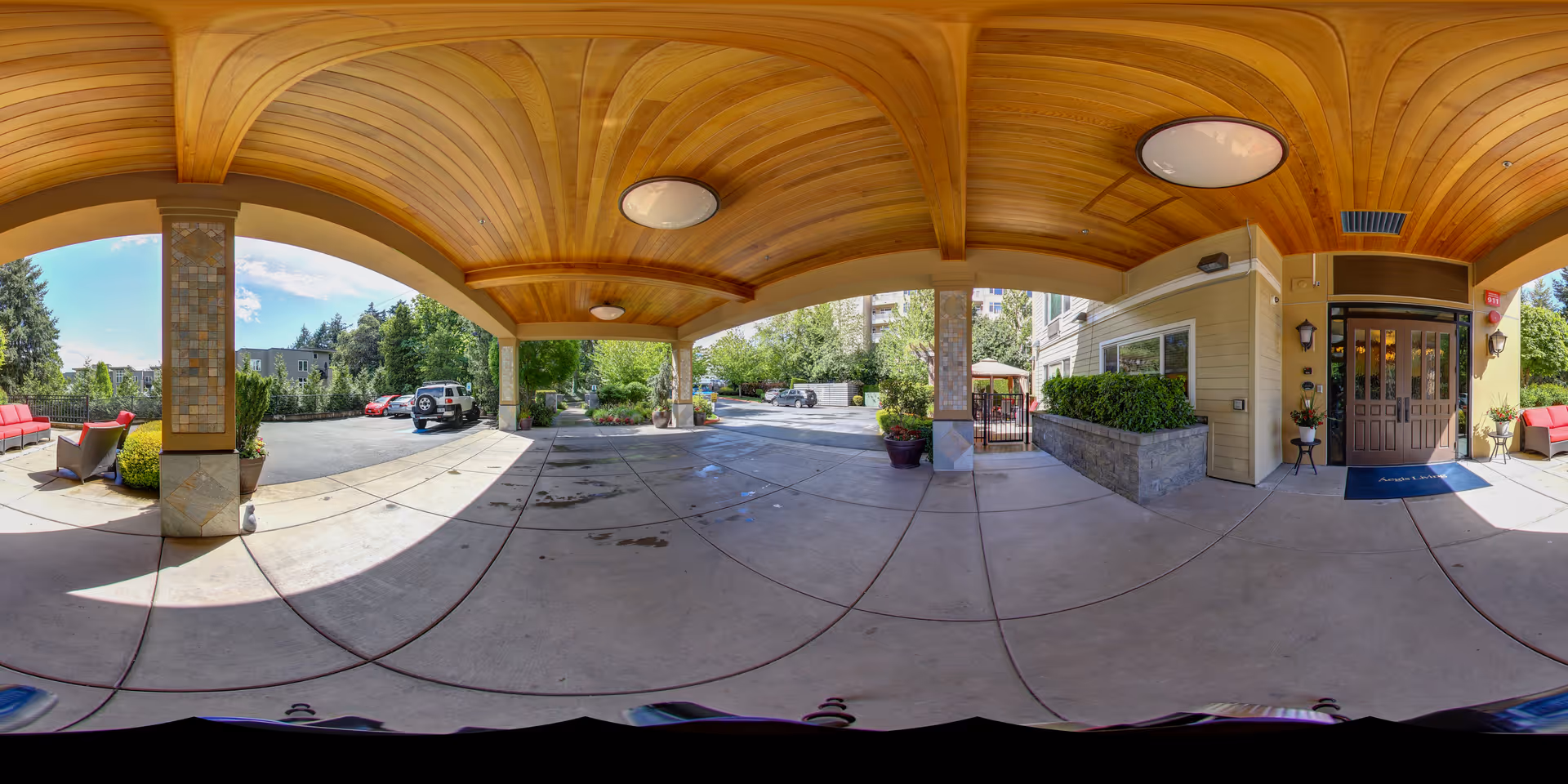 Covered porte-cochere entrance with a wooden arched ceiling, driveway, planters and seating in front of a senior living building.
