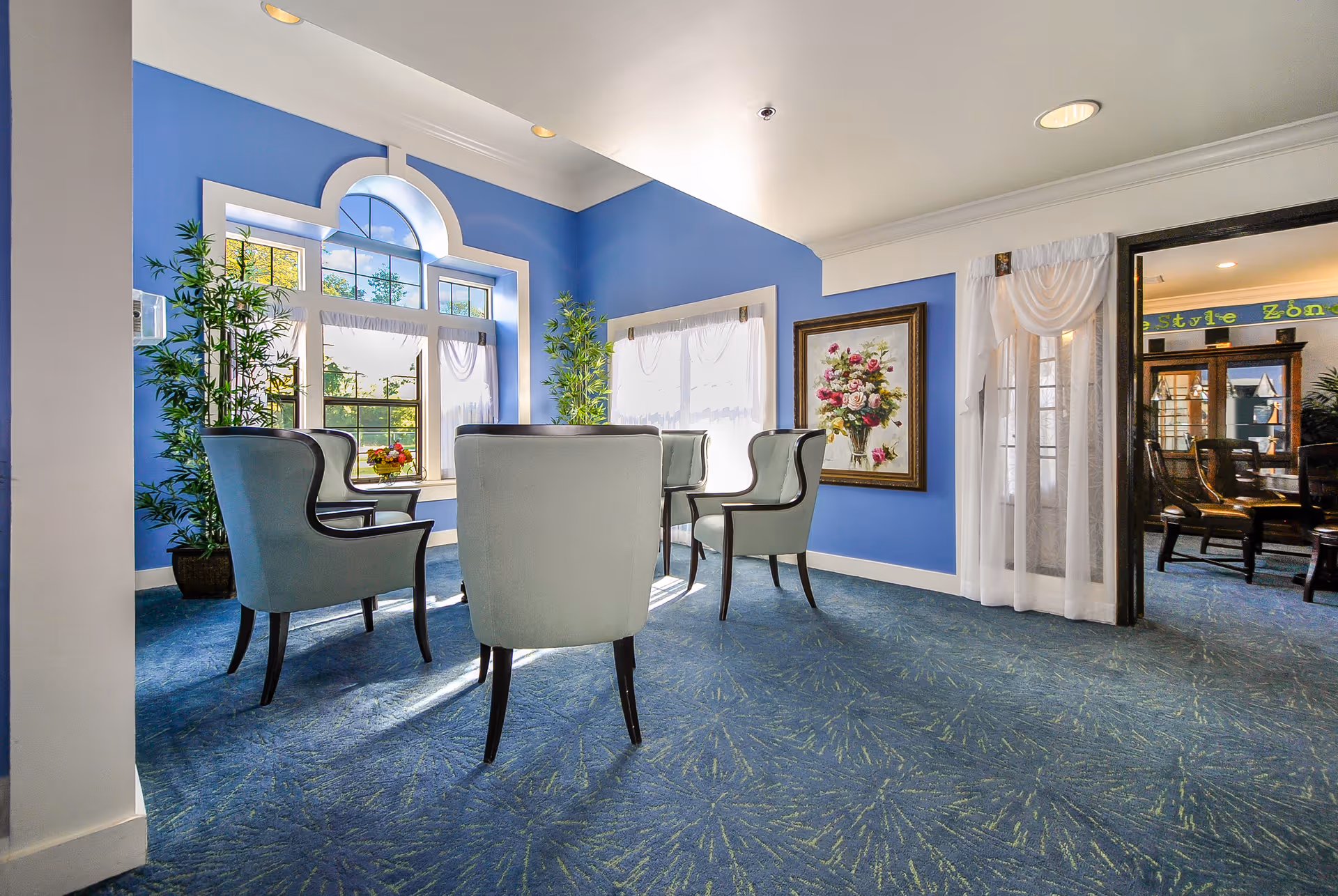 A bright sitting area with four light blue upholstered chairs arranged around a small table. The room has blue walls and carpet with a floral pattern. Large windows with white curtains allow natural light to fill the space. There are two tall potted plants near the windows and a framed floral painting on the wall. A doorway with sheer white curtains leads to another room with wooden chairs and a sign that reads 'Lifestyle Zone'.