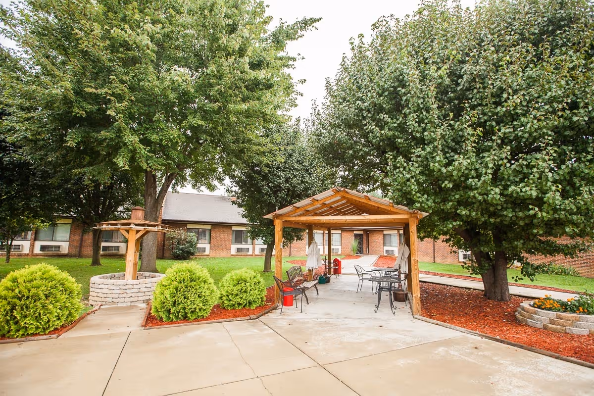 Outdoor courtyard with a wooden pergola, patio tables and chairs, trees, shrubs, and a surrounding brick building.