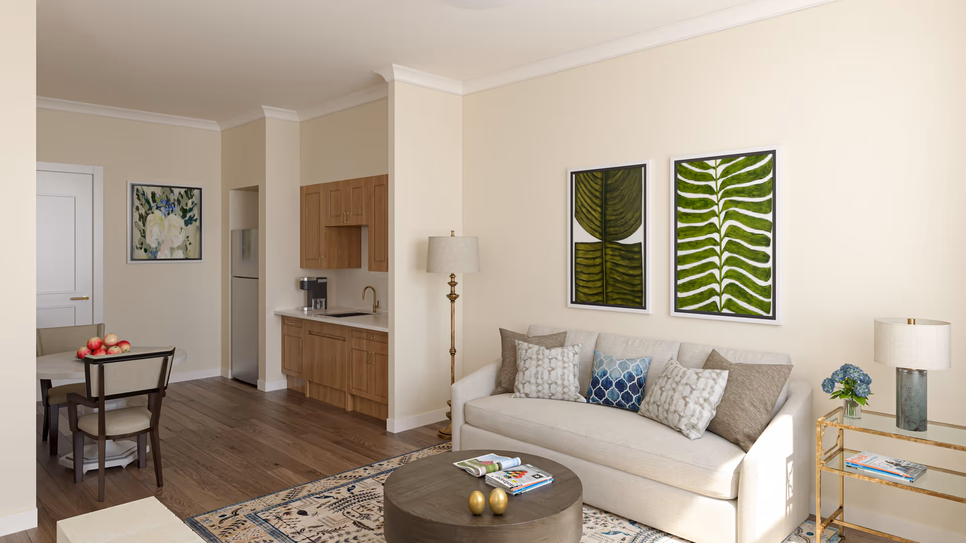 A cozy living room and kitchen area with beige walls and wooden flooring. The living room features a light beige sofa with multiple decorative pillows, a round wooden coffee table with magazines and decorative items, and a floor lamp. Two framed green leaf artworks hang above the sofa. To the right, there is a glass and gold side table with a lamp and a small vase with blue flowers. The kitchen area has wooden cabinets, a countertop with a sink, and a stainless steel refrigerator. A small dining table with four chairs and a bowl of apples is visible near the kitchen. A floral painting hangs on the wall near a white door.