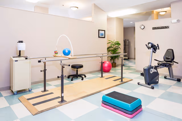 A physical therapy or rehabilitation room with parallel bars for walking exercises, a stationary exercise bike, a stack of aerobic step platforms, a small stool, a potted plant, and a small cabinet with a lamp. The room has a checkered blue and white floor and light-colored walls.