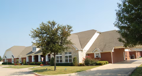 Exterior view of a single-story senior living facility building with a combination of brick and stone facade, multiple windows, a covered entrance, and a driveway. There are trees and well-maintained landscaping in front of the building under a clear blue sky.