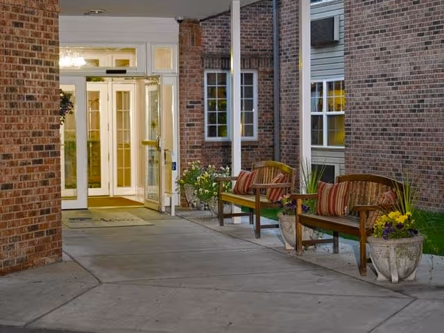 Covered outdoor entrance area of a brick building with two wooden benches, each with striped cushions, and large flower pots with plants on either side. The entrance has glass double doors with sidelights and transom windows, illuminated from inside.