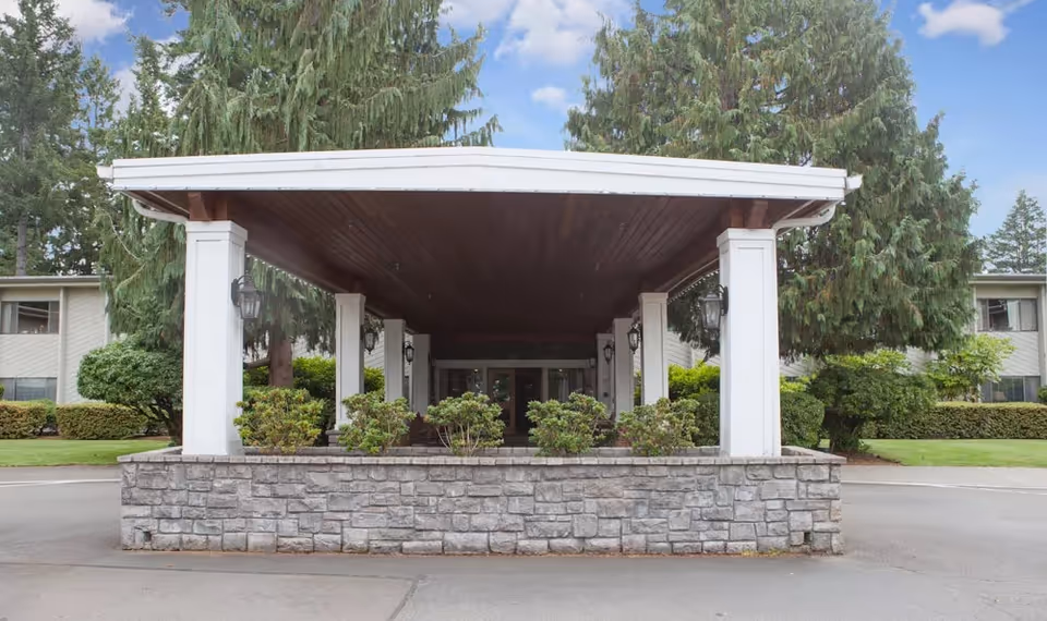 Covered entrance porte-cochère with white columns and a stone planter in front of a senior living building surrounded by trees.