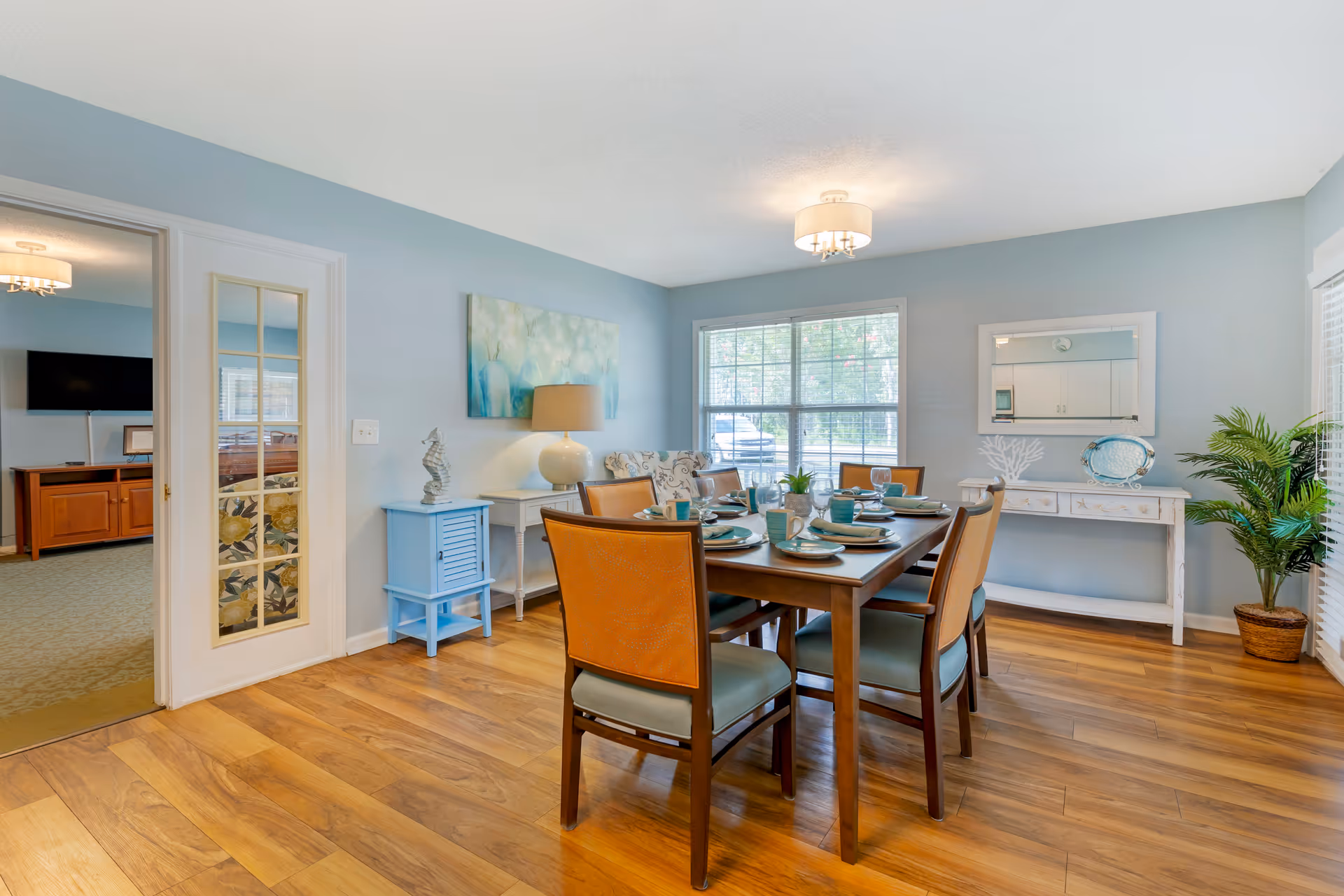 A bright dining room with a wooden table set for six people with plates, cups, and utensils. The room has light blue walls, a window letting in natural light, a white console table with decorative items, a potted plant, and a ceiling light fixture. A doorway with glass-paneled double doors leads to another room with a TV and wooden furniture.