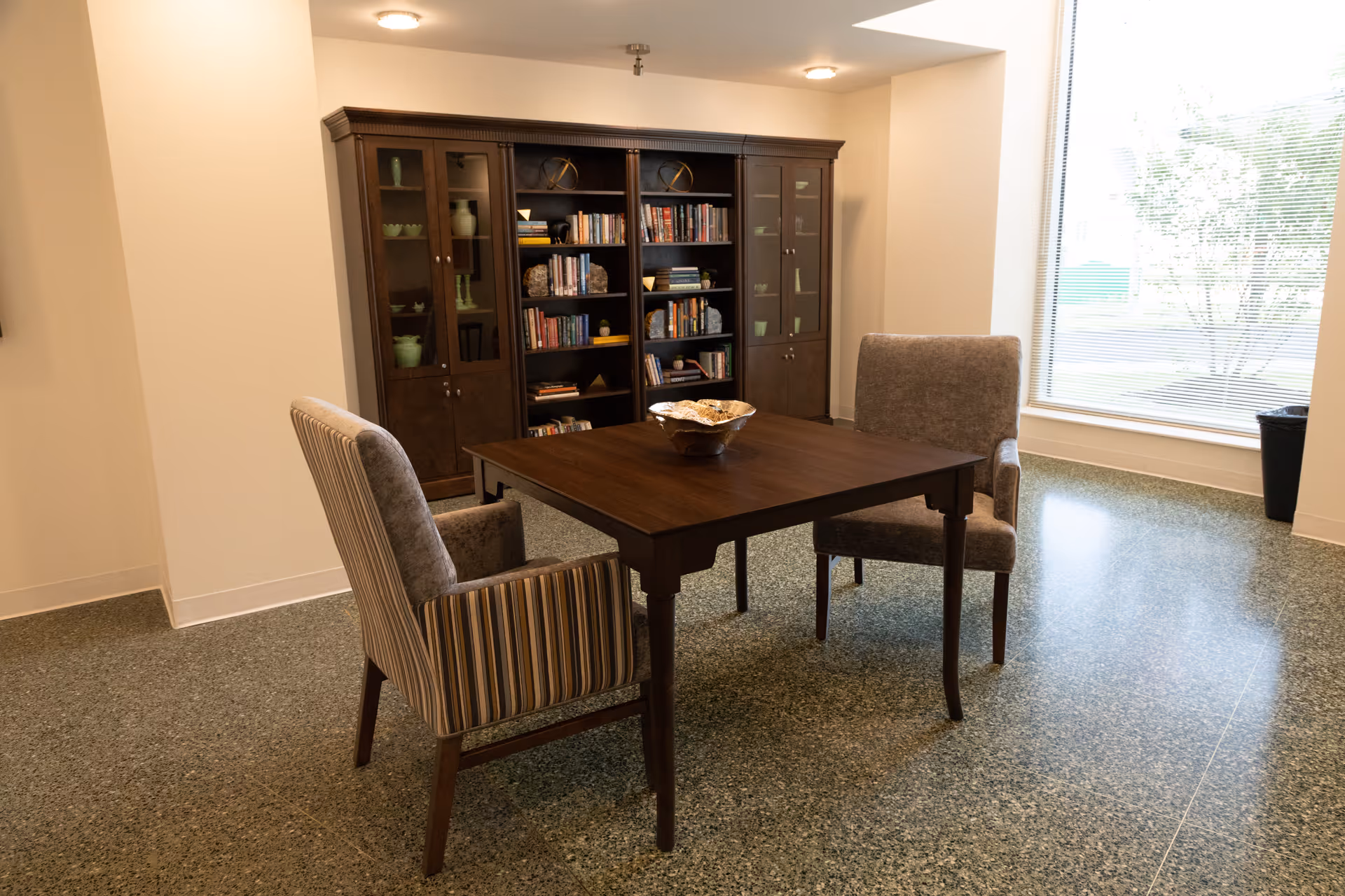 A quiet room with a wooden table and two upholstered chairs, one with striped fabric and the other in solid gray. Behind the table is a large wooden bookshelf filled with books and decorative items. A large window on the right side lets in natural light, illuminating the polished floor.