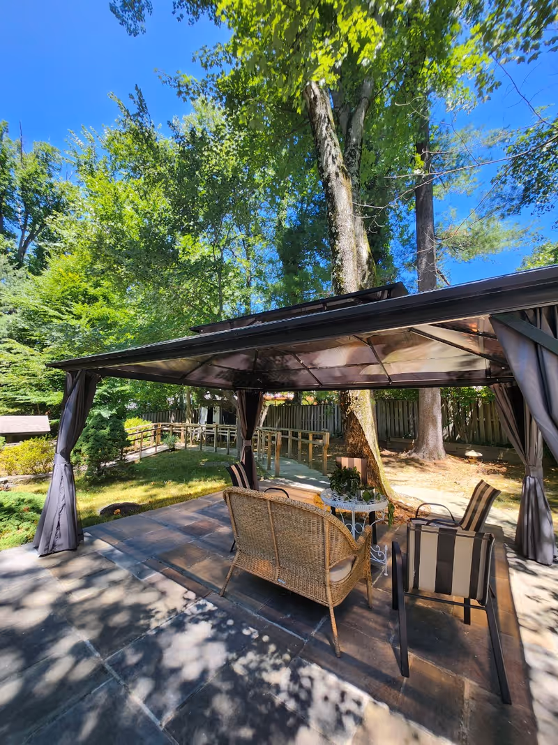 Outdoor patio area with a canopy providing shade over a wicker loveseat and two striped cushioned chairs around a small round table, surrounded by trees and greenery under a clear blue sky.