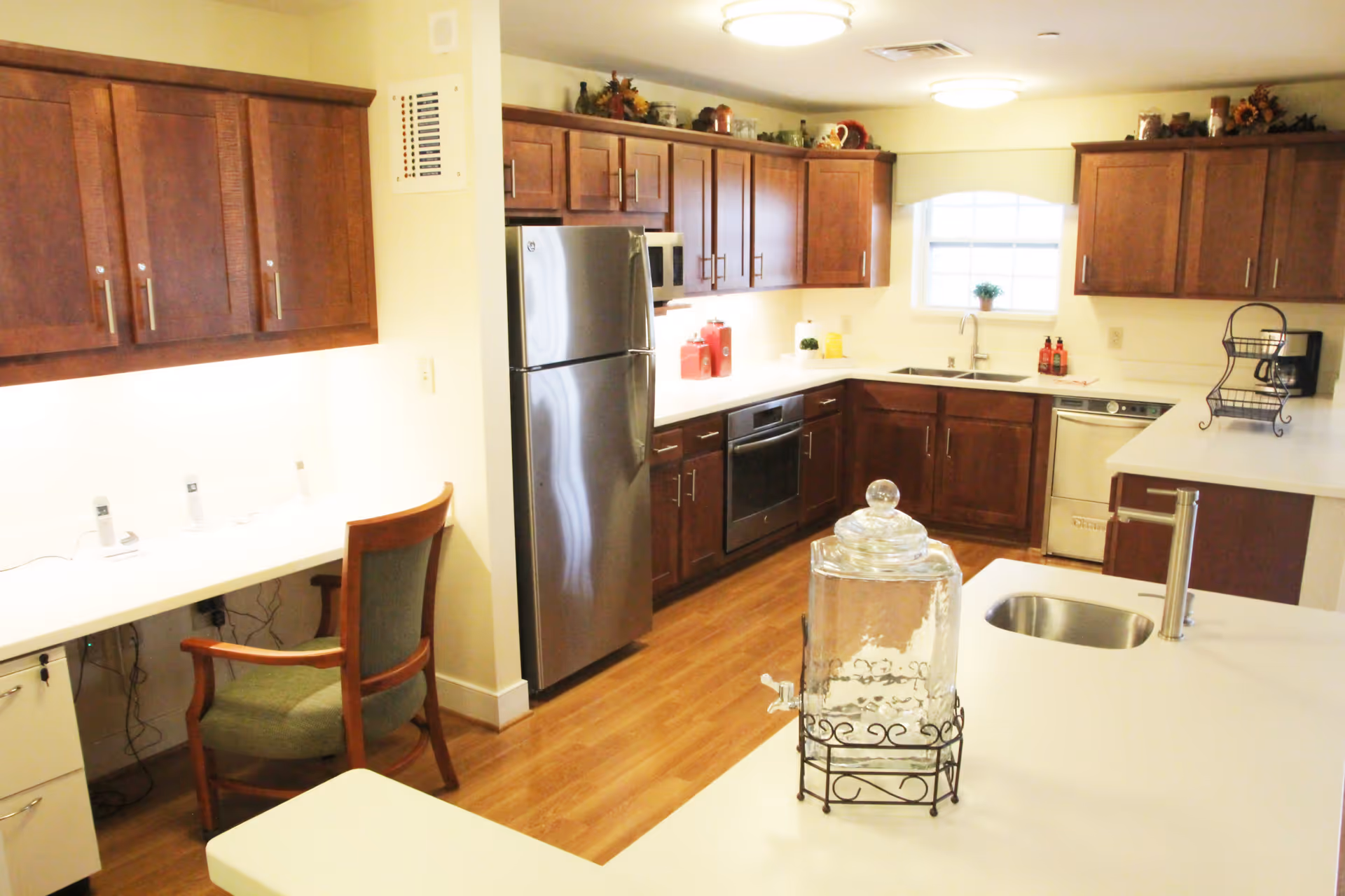 Bright kitchen area with wooden cabinets, stainless steel refrigerator, oven, dishwasher, and a sink. A glass water dispenser is placed on the white countertop island. There is a small desk area with a chair and some electronic devices on the left side. The room has wooden flooring and a window above the sink.