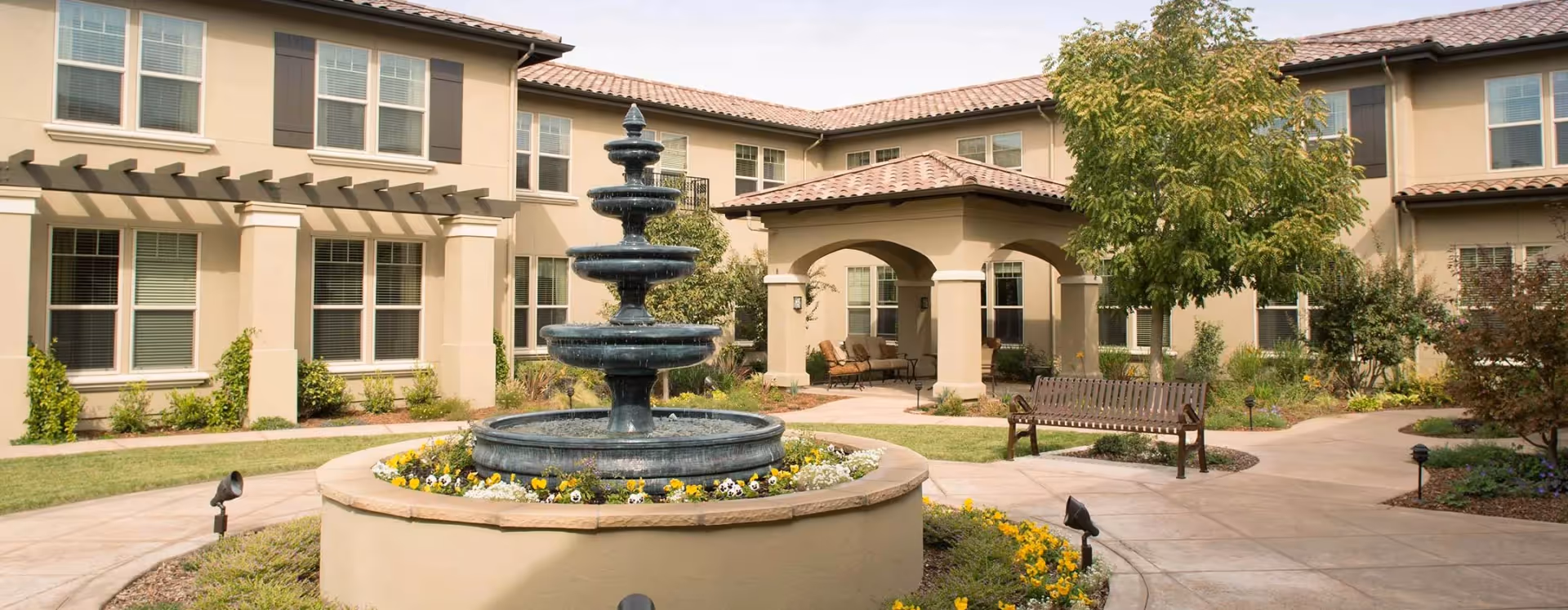 Courtyard of a senior living facility with a tiered fountain, flowerbeds, benches, and a surrounding two-story stucco building.