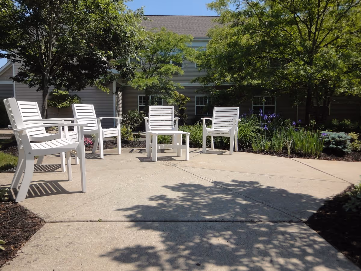 Outdoor patio area with white chairs and a small white table arranged on a concrete surface, surrounded by trees, plants, and flowers with a building visible in the background.