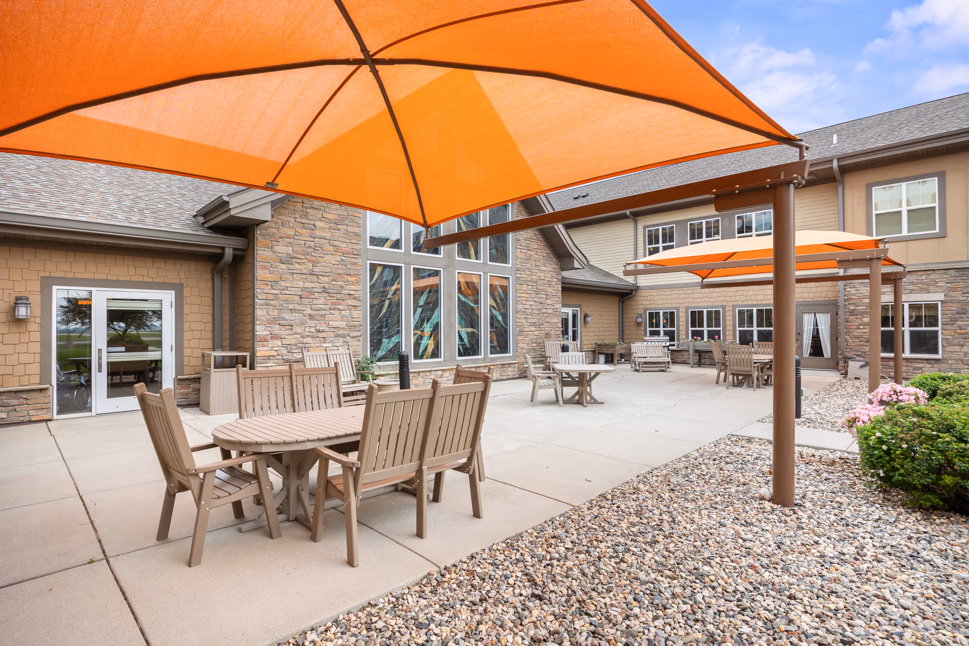 Outdoor patio courtyard with wooden tables and chairs under large orange umbrellas adjacent to a stone-and-siding building.