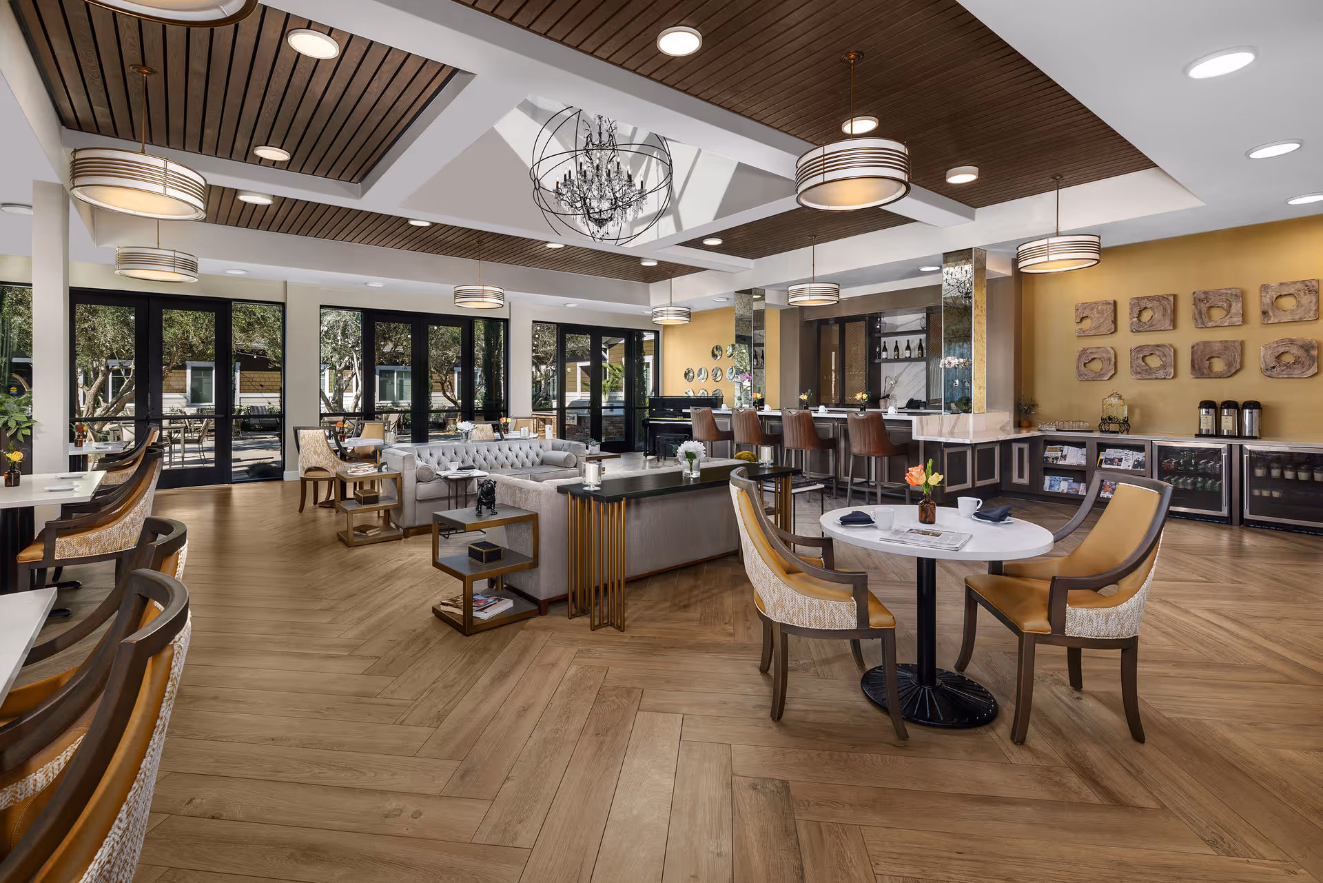Spacious and well-lit senior living facility common area with wooden flooring and ceiling accents, featuring a seating area with sofas and chairs, round tables with chairs, a bar counter with stools, decorative wall art, and large windows overlooking an outdoor patio.