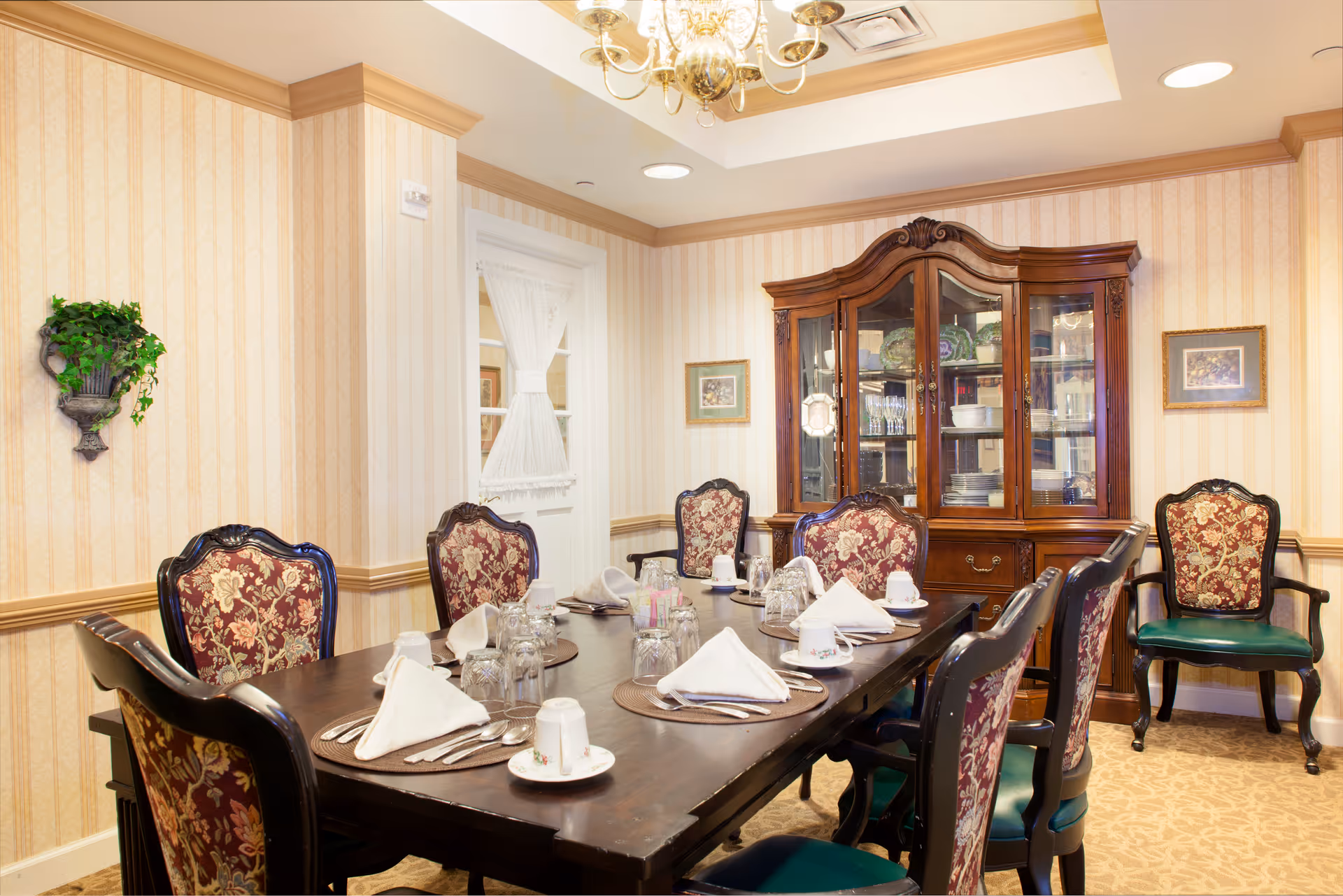 Elegant dining room with a long dark wooden table set with napkins, china and glassware, upholstered chairs, and a glass-front china cabinet against wallpapered walls.