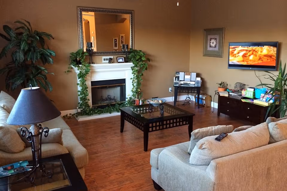 A cozy living room with two beige sofas, a glass-top coffee table, a fireplace decorated with green vines, a large mirror above the fireplace, a floor lamp, a TV mounted on the wall displaying a cooking show, and various plants and decorative items around the room.