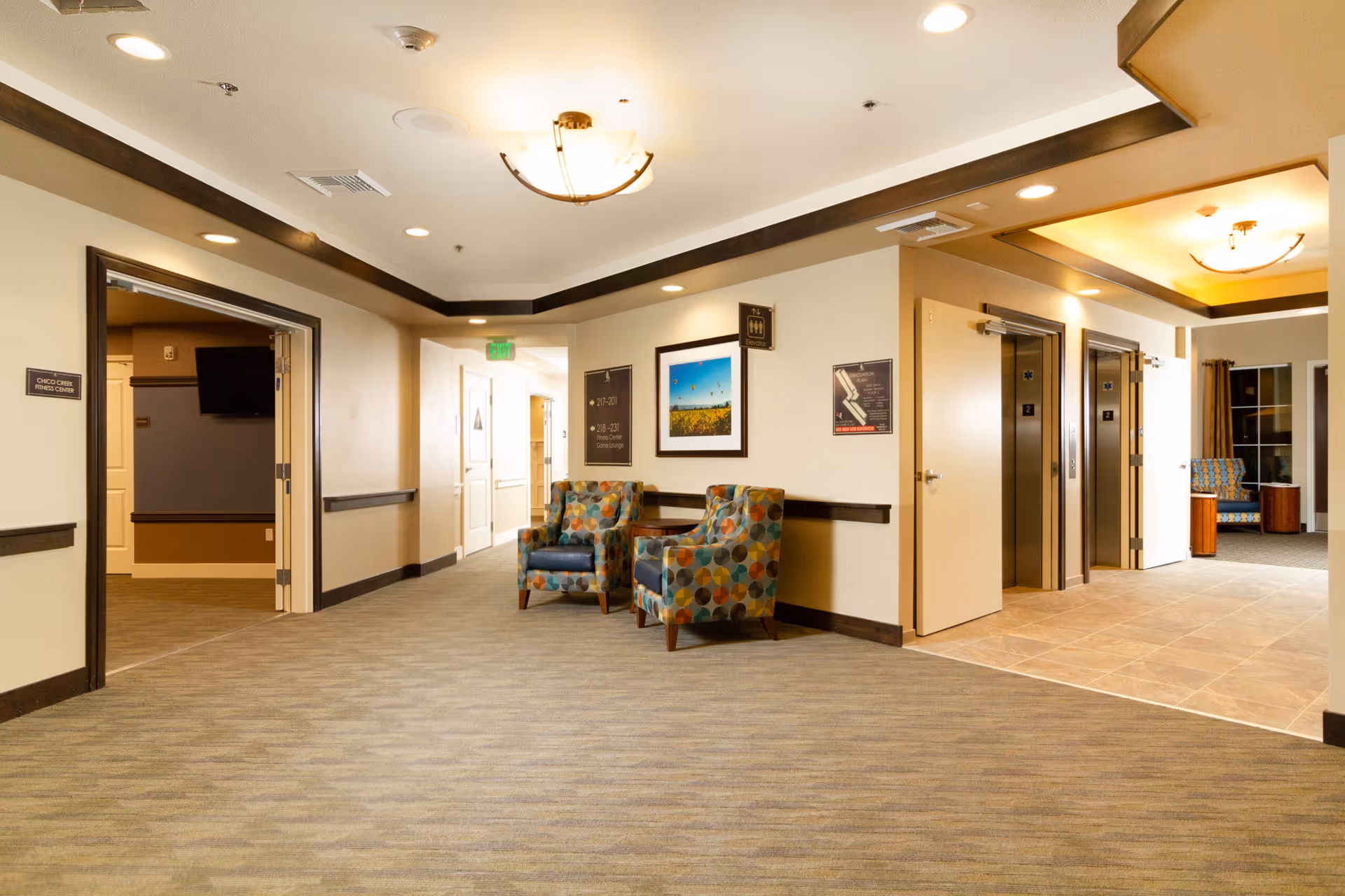 A well-lit hallway in a senior living facility featuring two patterned armchairs with a small table between them. The hallway has beige walls with dark wood trim, carpeted flooring, and ceiling lights. There are two elevators on the right side and an open doorway on the left leading to the Chico Creek Fitness Center. Signs on the walls indicate room numbers and directions.