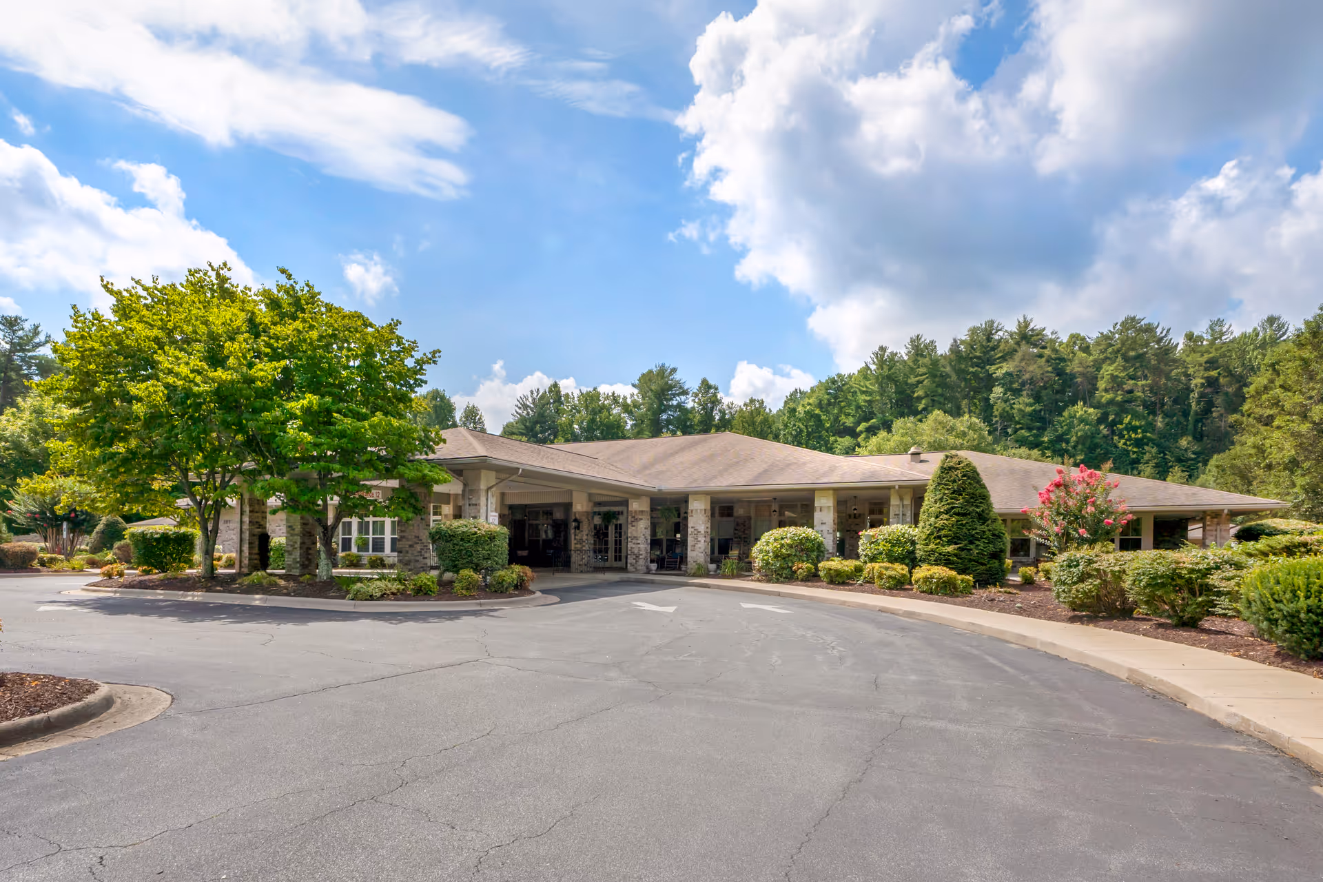 Exterior view of a single-story senior living facility building with a covered entrance, surrounded by landscaped bushes, trees, and a paved driveway under a partly cloudy sky.