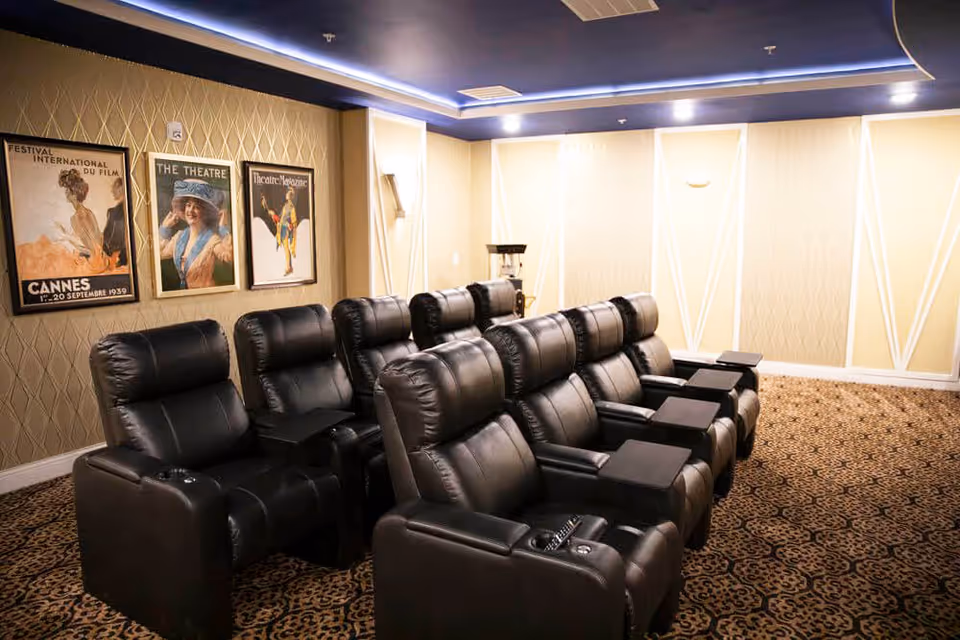 A small theater room with two rows of black leather recliner chairs, each equipped with armrests and cup holders. The walls are decorated with vintage movie posters, and the ceiling has recessed lighting with a blue accent. The carpet has an intricate pattern, and a popcorn machine is visible in the back corner.