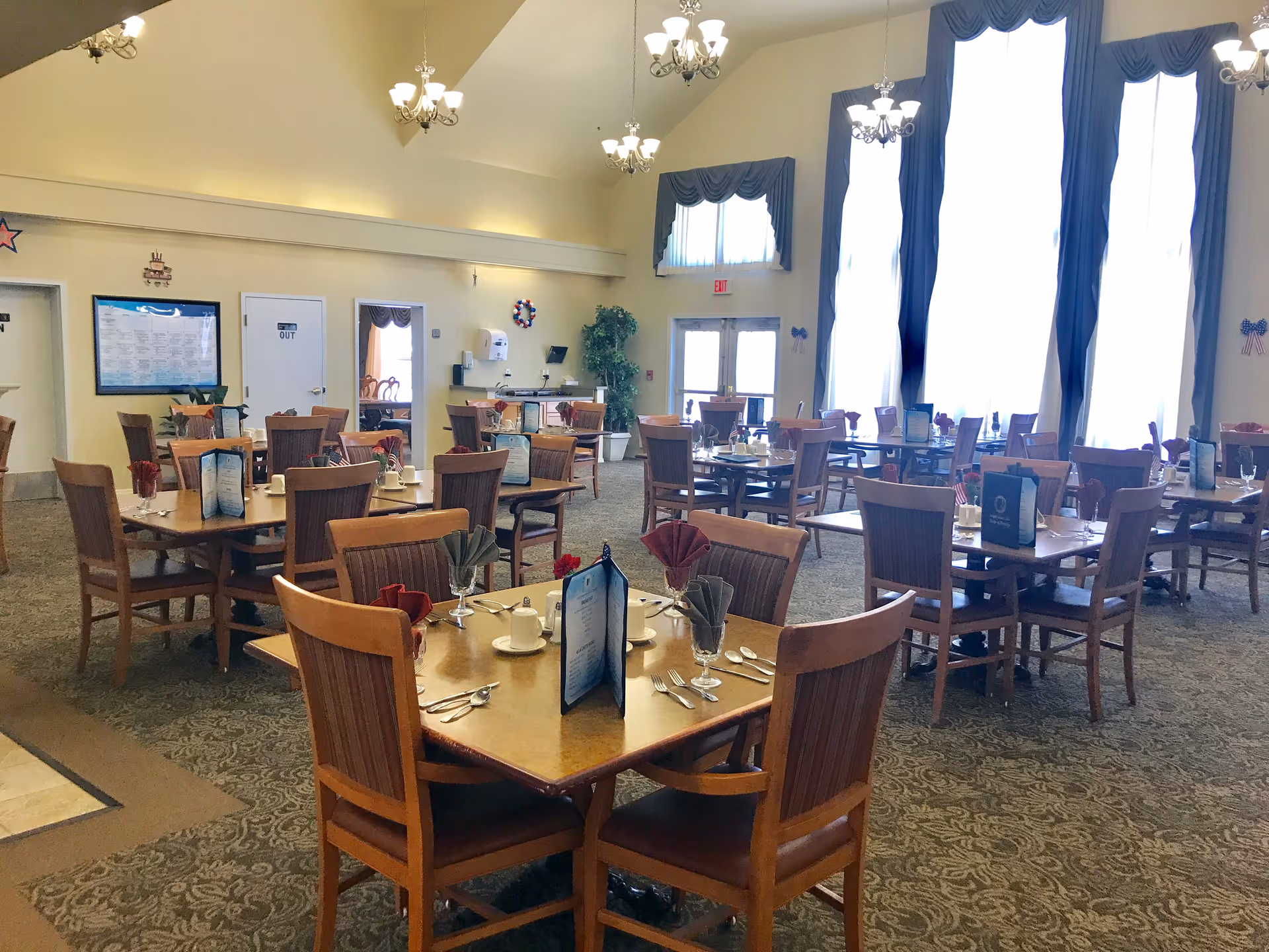 A spacious dining room with multiple wooden tables and chairs arranged neatly. Each table is set with menus, napkins folded in glasses, cups, and silverware. Large windows with blue curtains allow natural light to fill the room. Chandeliers hang from the high ceiling, and there is a carpeted floor with a patterned design.