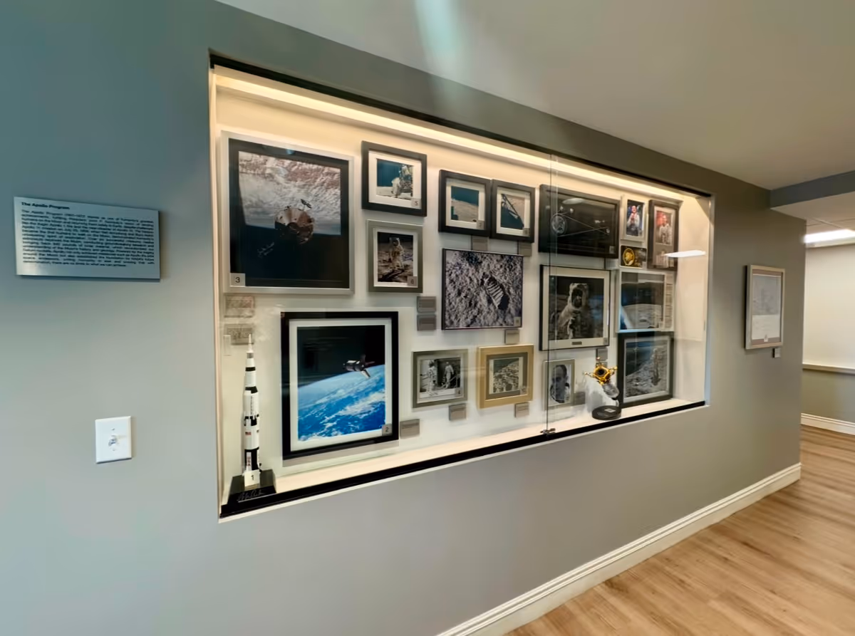 A hallway wall display featuring framed photographs and memorabilia related to the Apollo space program, including images of astronauts, the lunar surface, and a model rocket. The display is encased in glass with lighting, and there is a descriptive plaque on the left side of the display. The hallway has light-colored walls and wood flooring.
