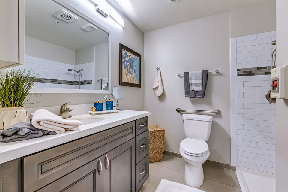 A clean and modern bathroom featuring a large mirror above a sink with a gray wooden cabinet. On the countertop are folded towels, a small plant, and bathroom accessories. The bathroom includes a white toilet with a grab bar beside it, a wicker laundry basket, and a walk-in shower with white subway tiles and a decorative tile strip. Towels hang on wall-mounted racks, and a framed artwork is displayed on the wall.