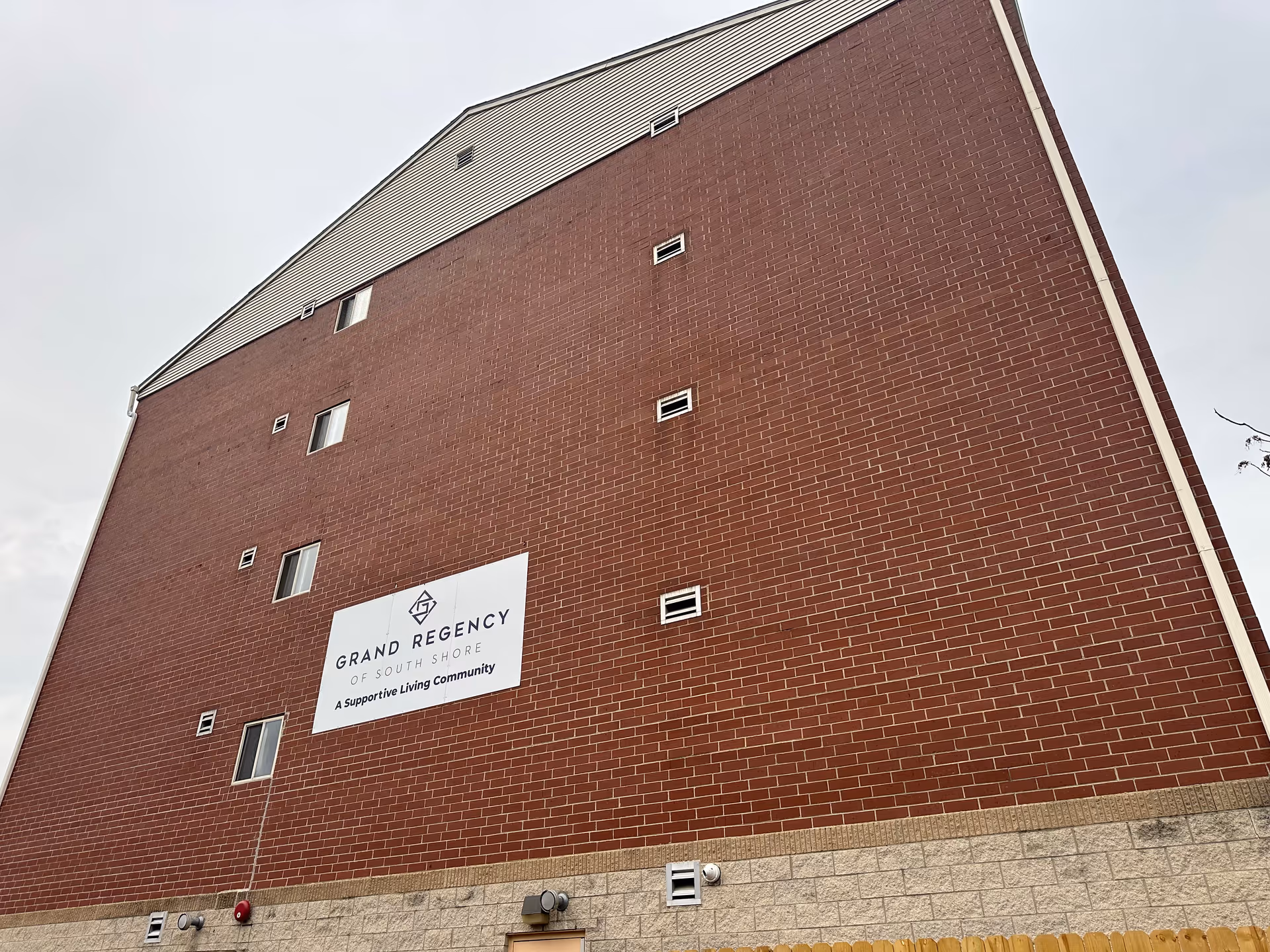 Exterior view of a multi-story brick building with a sign that reads 'Grand Regency of South Shore, A Supportive Living Community' on the wall under a cloudy sky.