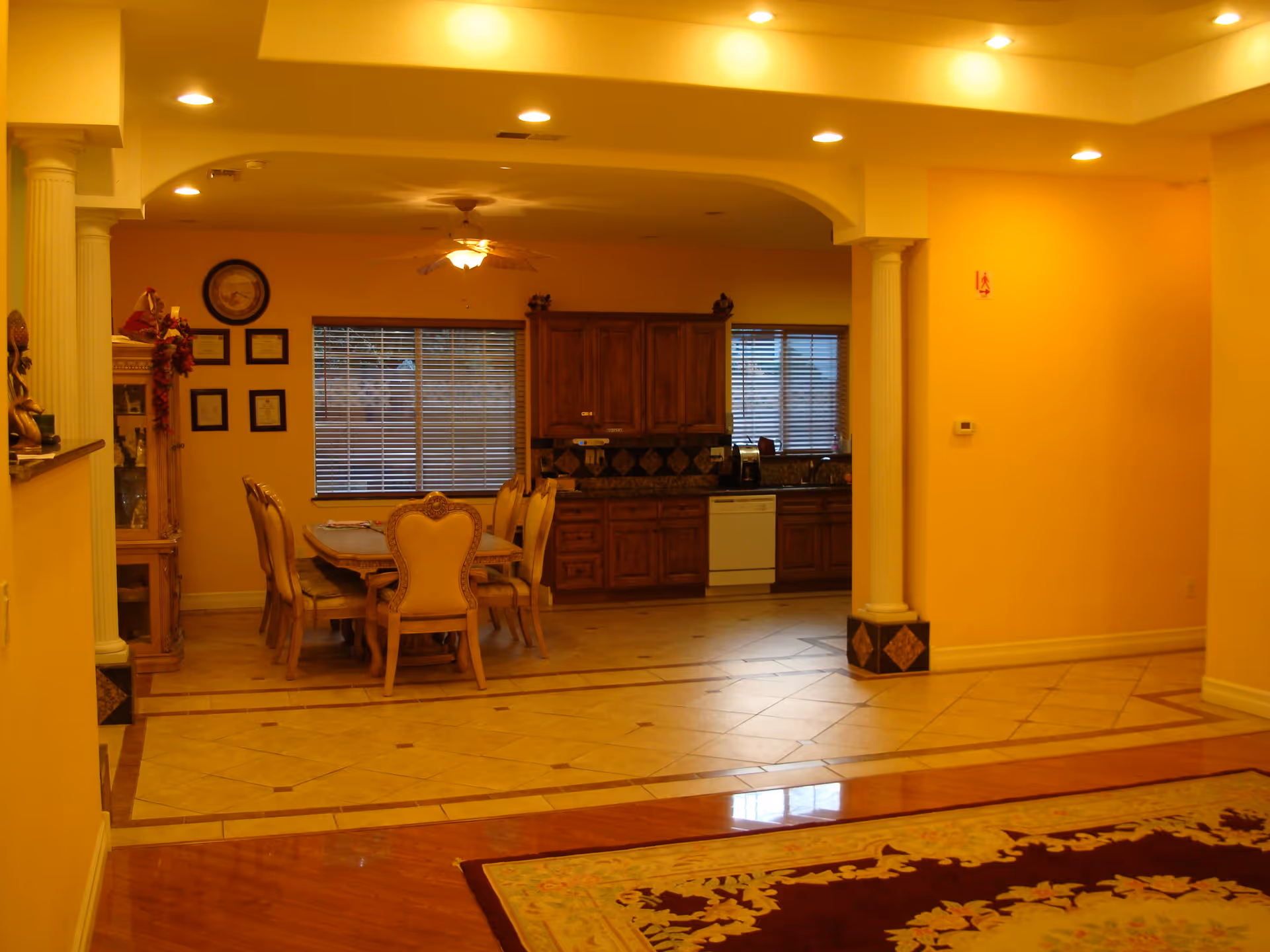 Interior view of a dining area and kitchen in a senior living facility. The dining area features a wooden table with six ornate chairs. The kitchen has wooden cabinets, a dishwasher, and windows with blinds. The room is warmly lit with recessed ceiling lights and a ceiling fan above the dining table. The floor transitions from tiled in the kitchen and dining area to wood in the foreground, with a decorative rug partially visible.