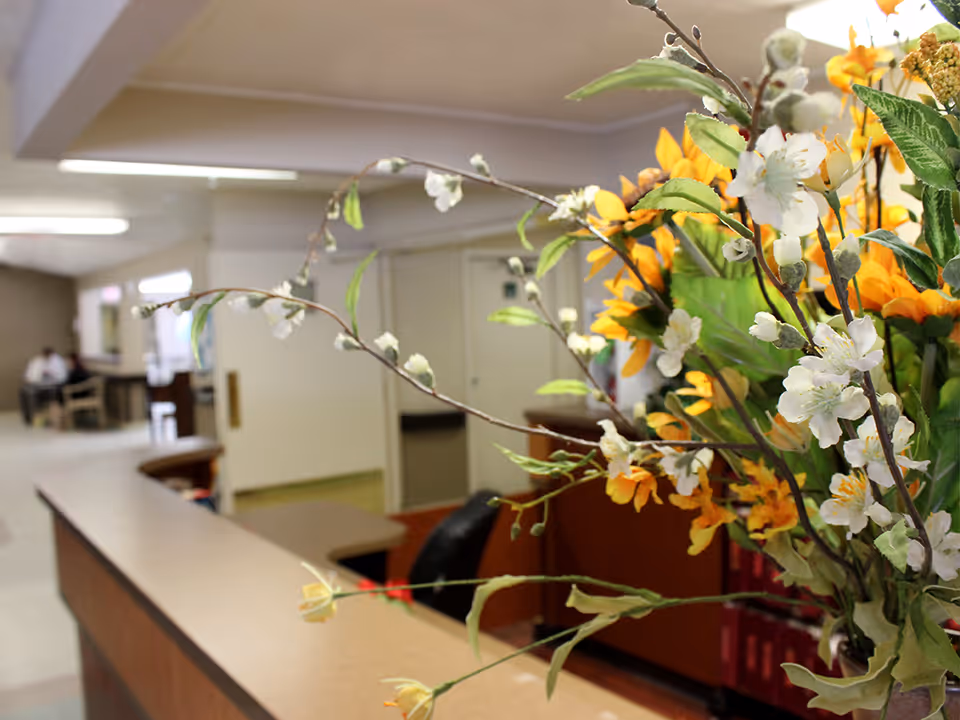 Close-up of a floral arrangement with white and yellow flowers on a reception desk in a senior living facility. In the background, there is a hallway with a person sitting at a table and doors along the corridor.