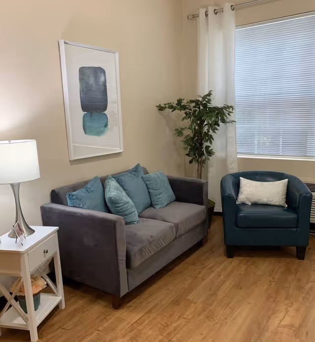 A cozy living room area with a gray sofa adorned with four teal and blue cushions, a teal armchair with a white pillow, a white side table with a lamp and a small plant, a framed abstract artwork on the wall, and a window with white curtains and blinds letting in natural light.