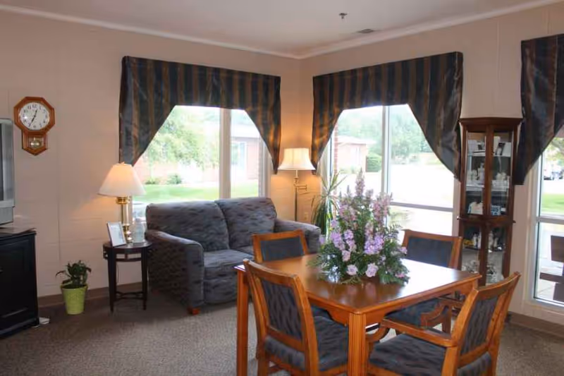 A cozy living room area with a wooden dining table surrounded by four chairs, a gray upholstered loveseat, two table lamps, a wall clock, a small side table with a photo frame, a green potted plant, and a glass display cabinet. Large windows with striped valances let in natural light and show a view of greenery outside.