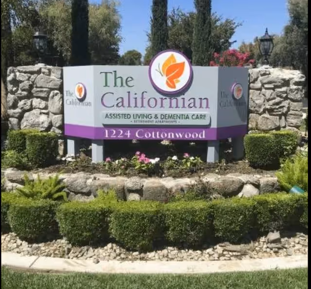 Outdoor stone sign for The Californian assisted living and dementia care facility, surrounded by neatly trimmed bushes and flowers, with trees and a clear blue sky in the background.