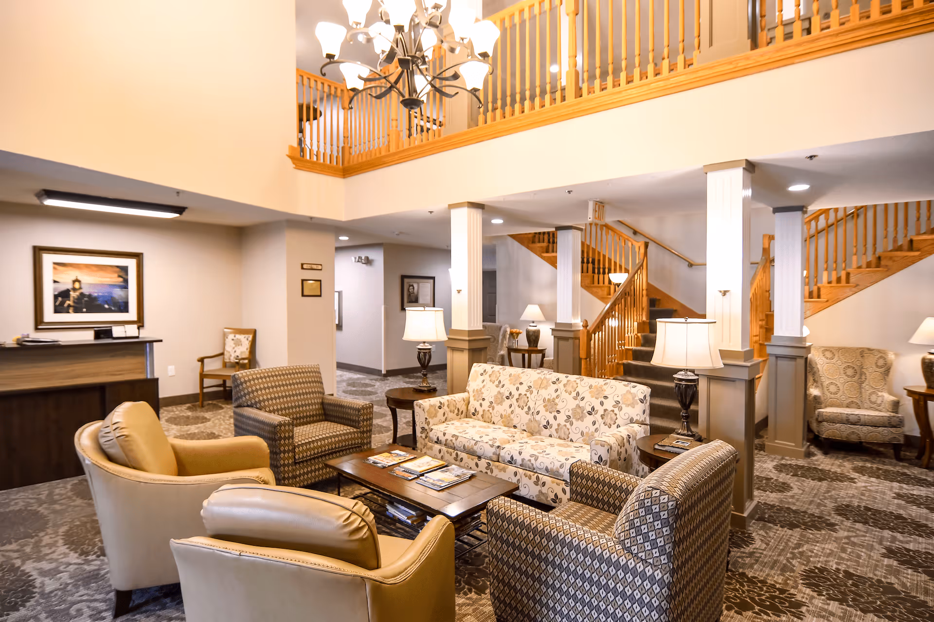 A spacious senior living facility common area with comfortable seating including a floral-patterned sofa, two patterned armchairs, and two beige armchairs arranged around a wooden coffee table. The room features carpeted floors, a wooden staircase with railings leading to an upper level, several table lamps, and a chandelier hanging from the ceiling. There is a reception desk and framed artwork on the walls.