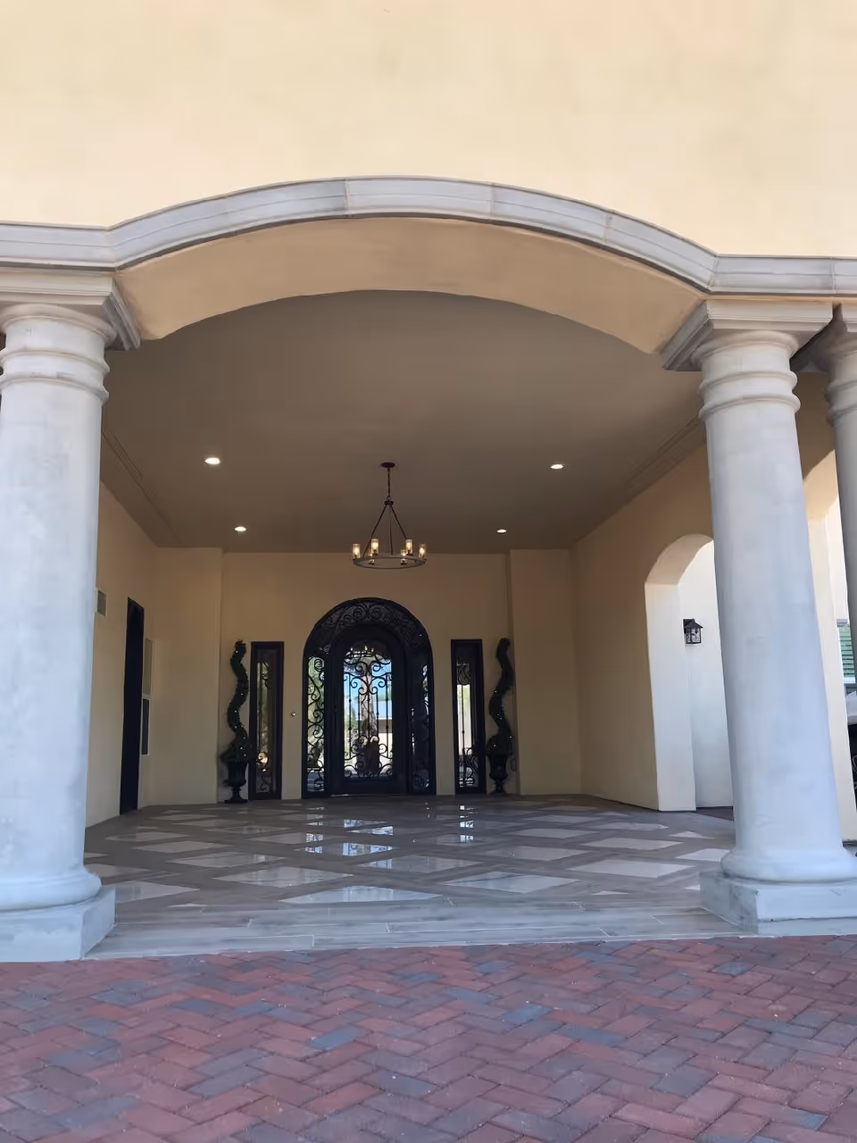 Covered entrance area with two large white columns supporting an archway. The floor has a checkered tile pattern, and there is a decorative black wrought iron door with glass panels at the back. Two topiary plants in black urns flank the door, and a chandelier with candles hangs from the ceiling.