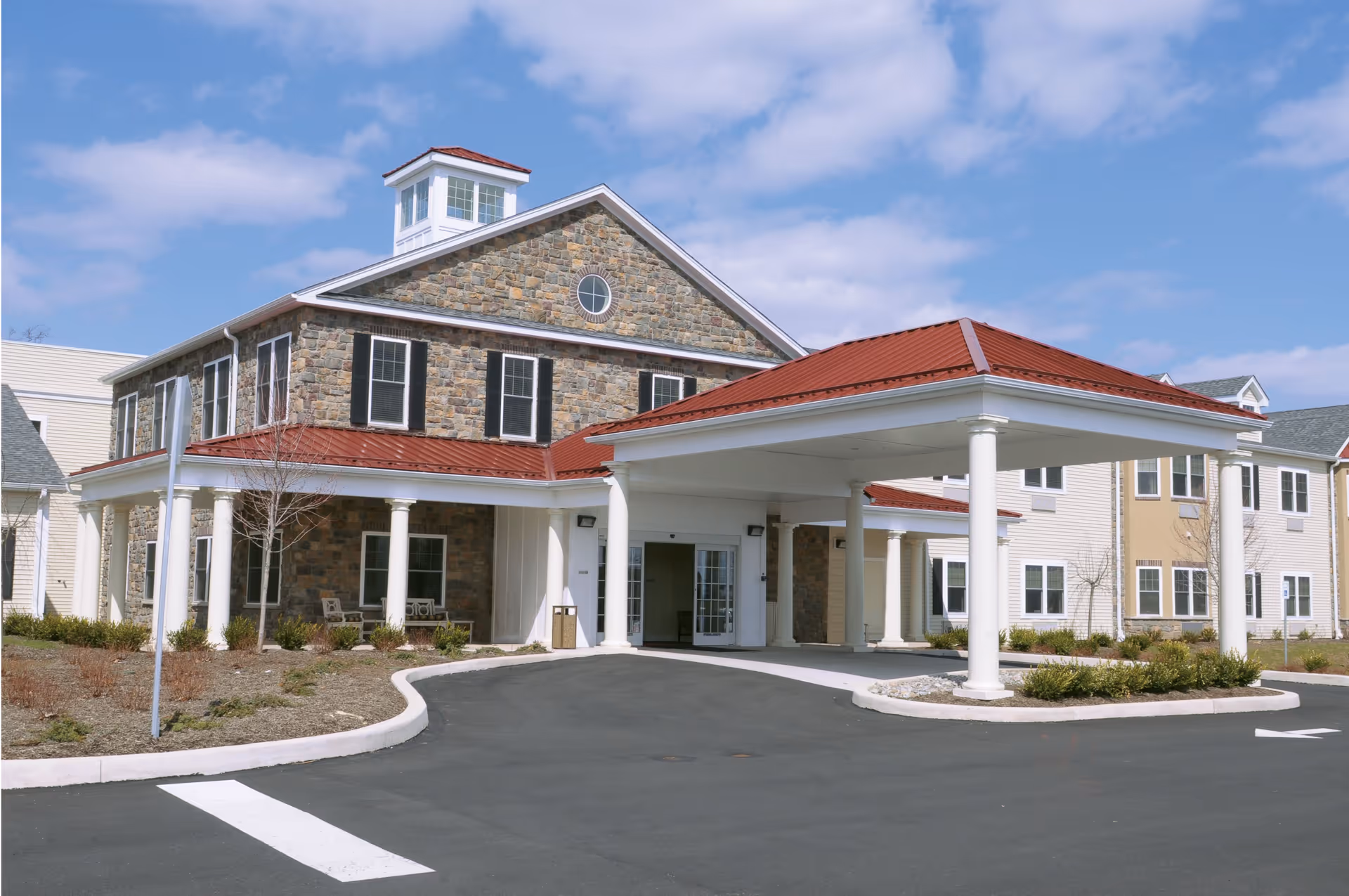 Exterior view of a senior living facility building with stone and siding walls, a red metal roof, white columns supporting a covered entrance, and a paved driveway under a partly cloudy sky.