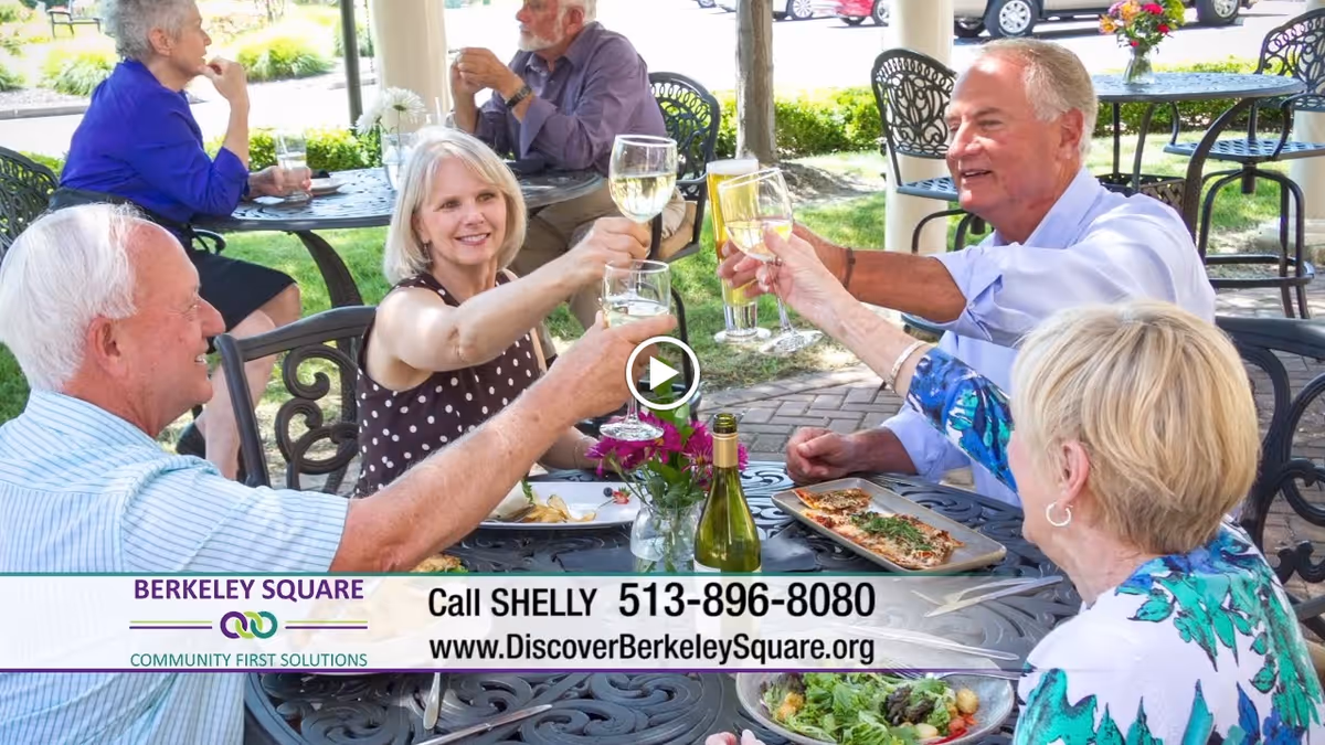 A group of elderly people sitting outdoors at a round metal table, enjoying food and drinks while toasting with wine glasses. The setting is a patio area with greenery and parked cars in the background.