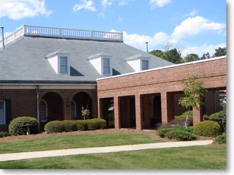 Exterior view of a brick building with a gray roof featuring three dormer windows and a white railing on top. The building has an arched entrance and a covered walkway supported by brick columns. There is a well-maintained lawn with shrubs and small trees in front of the building under a partly cloudy blue sky.