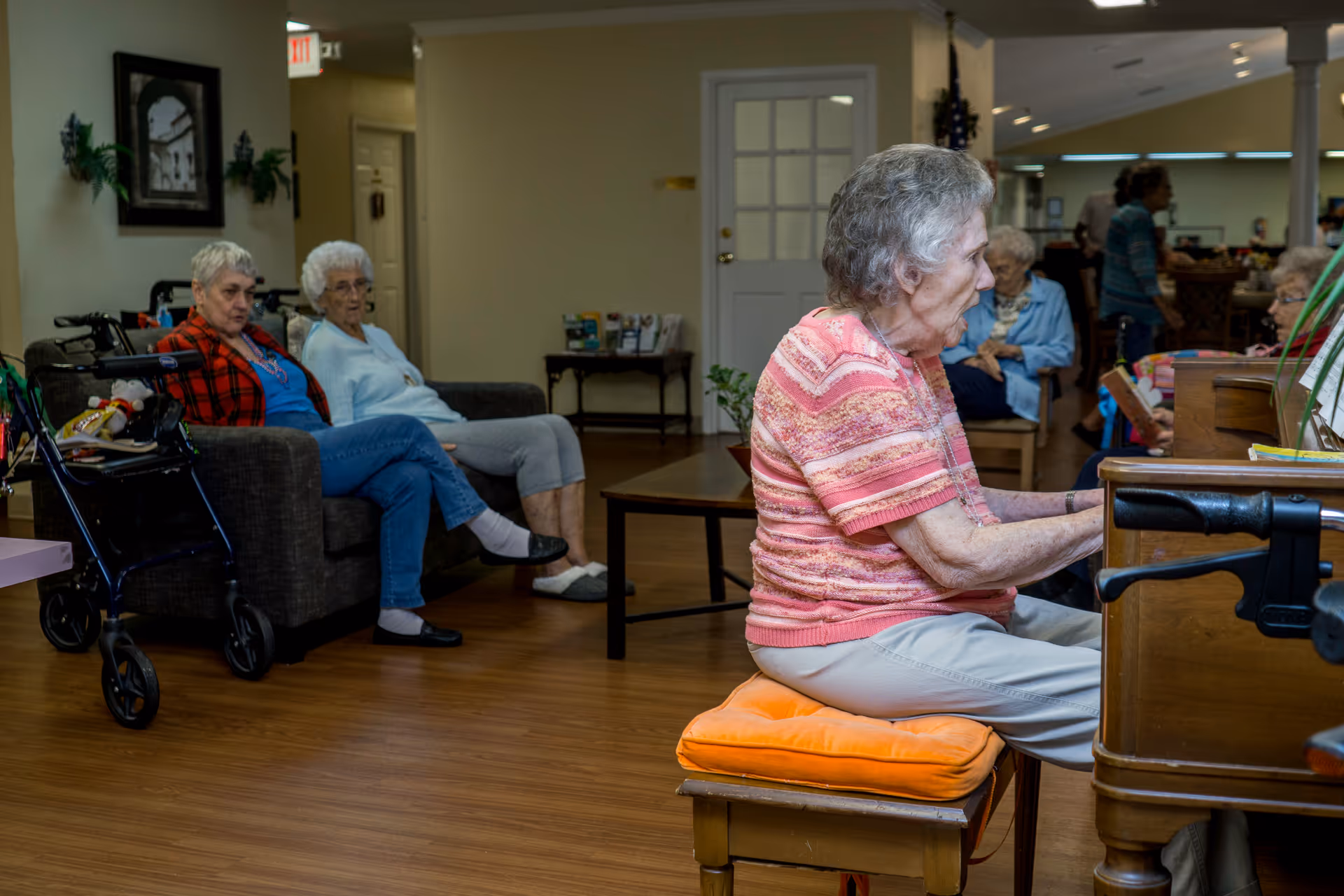 Several elderly residents sit and socialize in a senior living common room while a woman plays the piano.