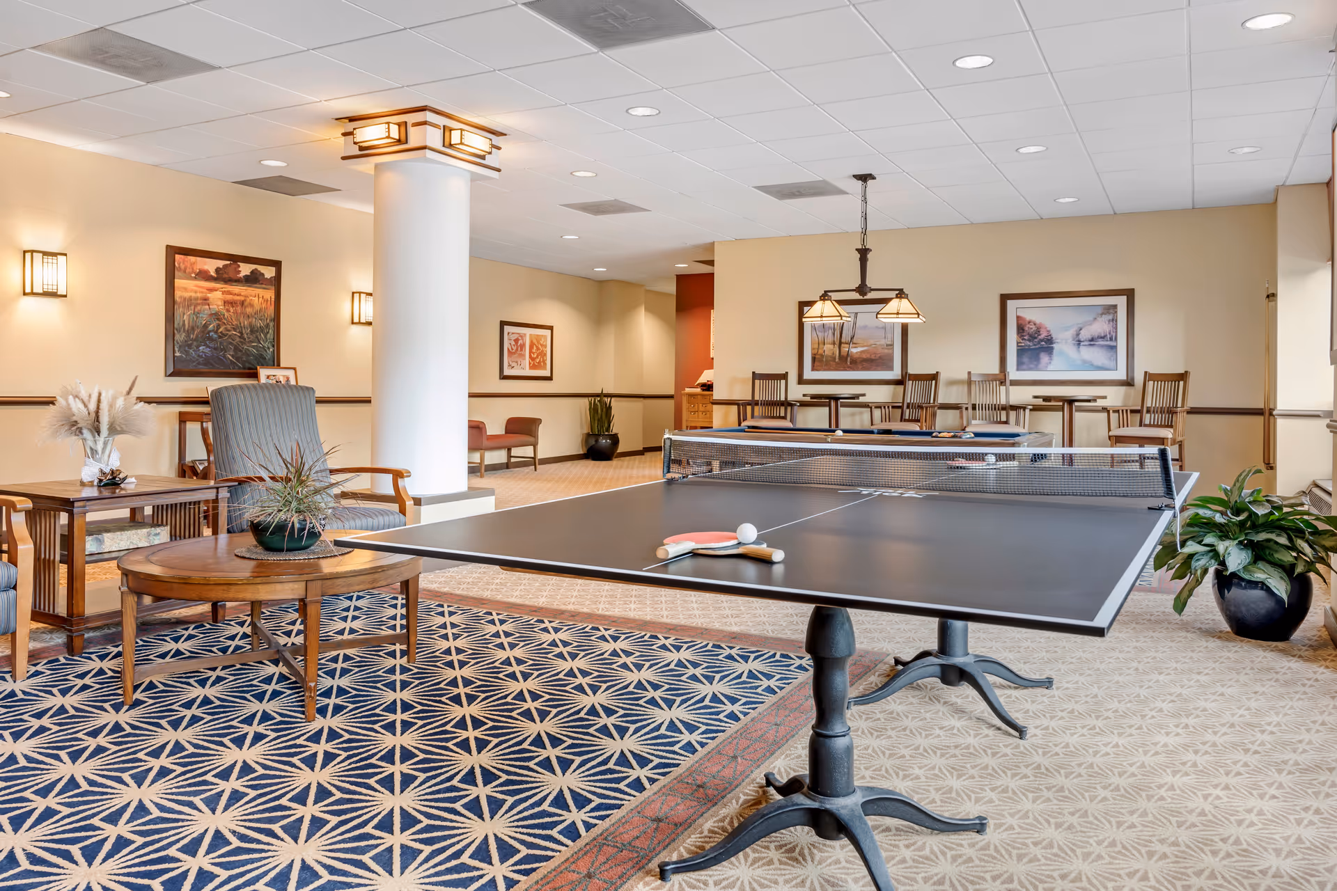 A spacious recreational room with a ping pong table in the foreground, two paddles and a ball on the table. The room features comfortable seating including armchairs and a round wooden table with plants on it. The walls are decorated with framed landscape paintings and the ceiling has recessed lighting. There is a carpet with a geometric pattern and a large white column with a decorative light fixture. In the background, there are high tables with chairs and hanging lights.
