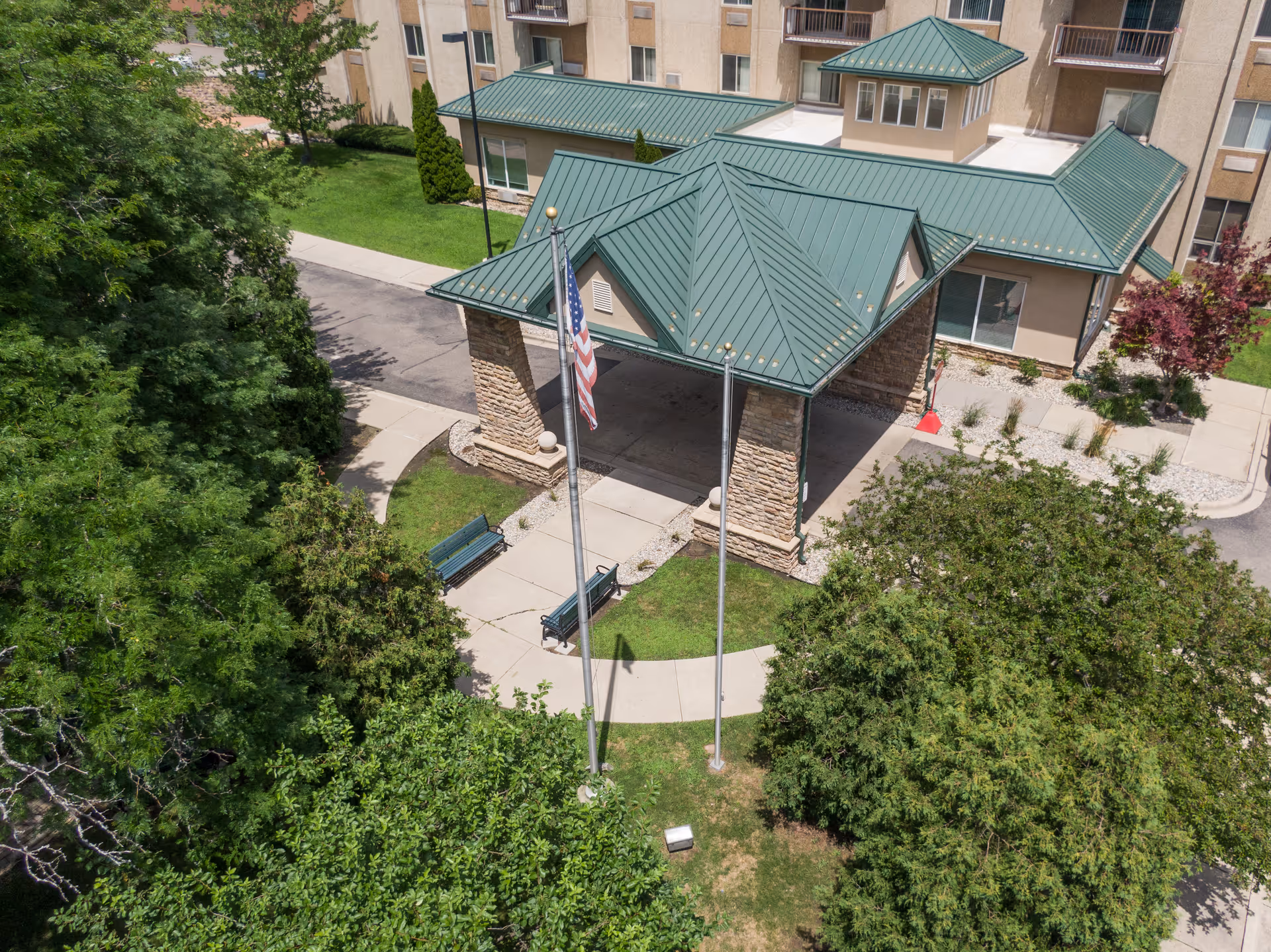Aerial view of the entrance area of a senior living facility with a green metal roof canopy supported by stone pillars. There are two flagpoles, one with an American flag, and two green benches on a concrete walkway surrounded by grass and trees. The building has beige walls and multiple windows.
