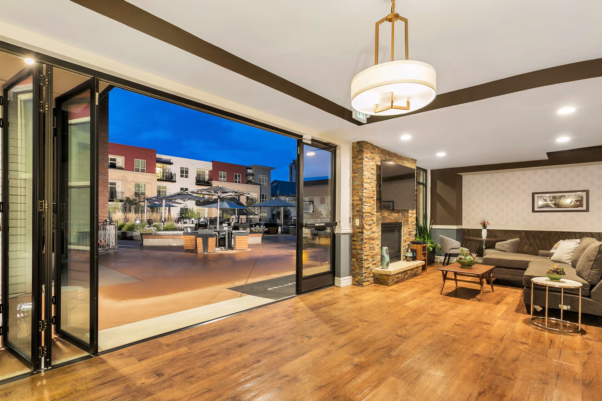 A spacious living room area with wooden flooring, a stone fireplace with a mounted TV above it, a gray sectional sofa, two gray chairs, and a coffee table. Large folding glass doors open to an outdoor patio area with tables, umbrellas, and surrounding apartment buildings under a twilight sky.