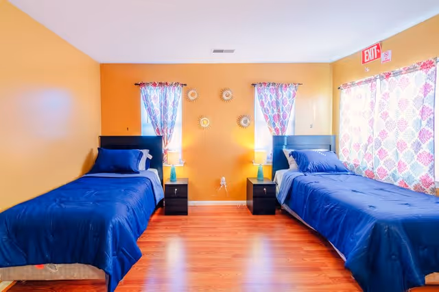 A bright bedroom with two twin beds, each with dark blue bedding and pillows. Between the beds are two black nightstands with matching lamps. The walls are painted orange, and there are two windows with colorful patterned curtains behind each bed. The floor is wooden, and there is an exit sign above a window on the right side.