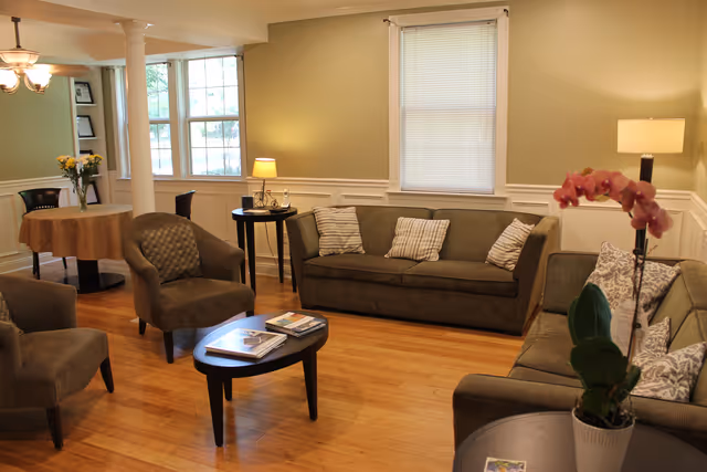 A cozy living room area in a memory care home with two sofas and two armchairs arranged around a small oval coffee table with magazines. There is a round dining table with chairs in the background near windows letting in natural light. The room has wooden flooring, neutral-colored walls, and is decorated with table lamps, cushions, and a potted orchid on a side table.
