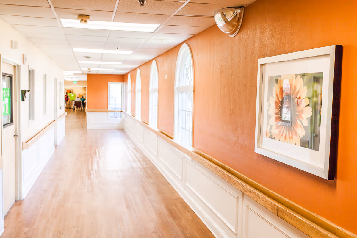 A long, bright hallway in a senior living facility with wooden floors, orange walls on one side with framed floral artwork, and white walls with windows on the other side. The hallway has handrails on both sides and a ceiling with recessed lighting. At the far end, there are people and a door visible.