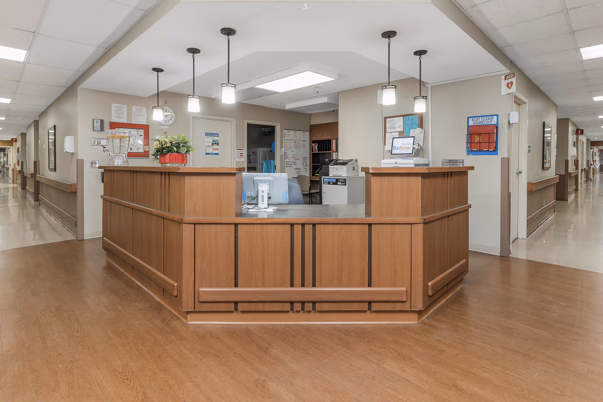 A reception desk area in a senior care facility with wooden paneling and a gray countertop. The desk has a computer monitor, a printer, a flower arrangement, and various office supplies. The area is well-lit with ceiling lights and pendant lamps. Hallways extend on both sides of the desk with handrails along the walls.